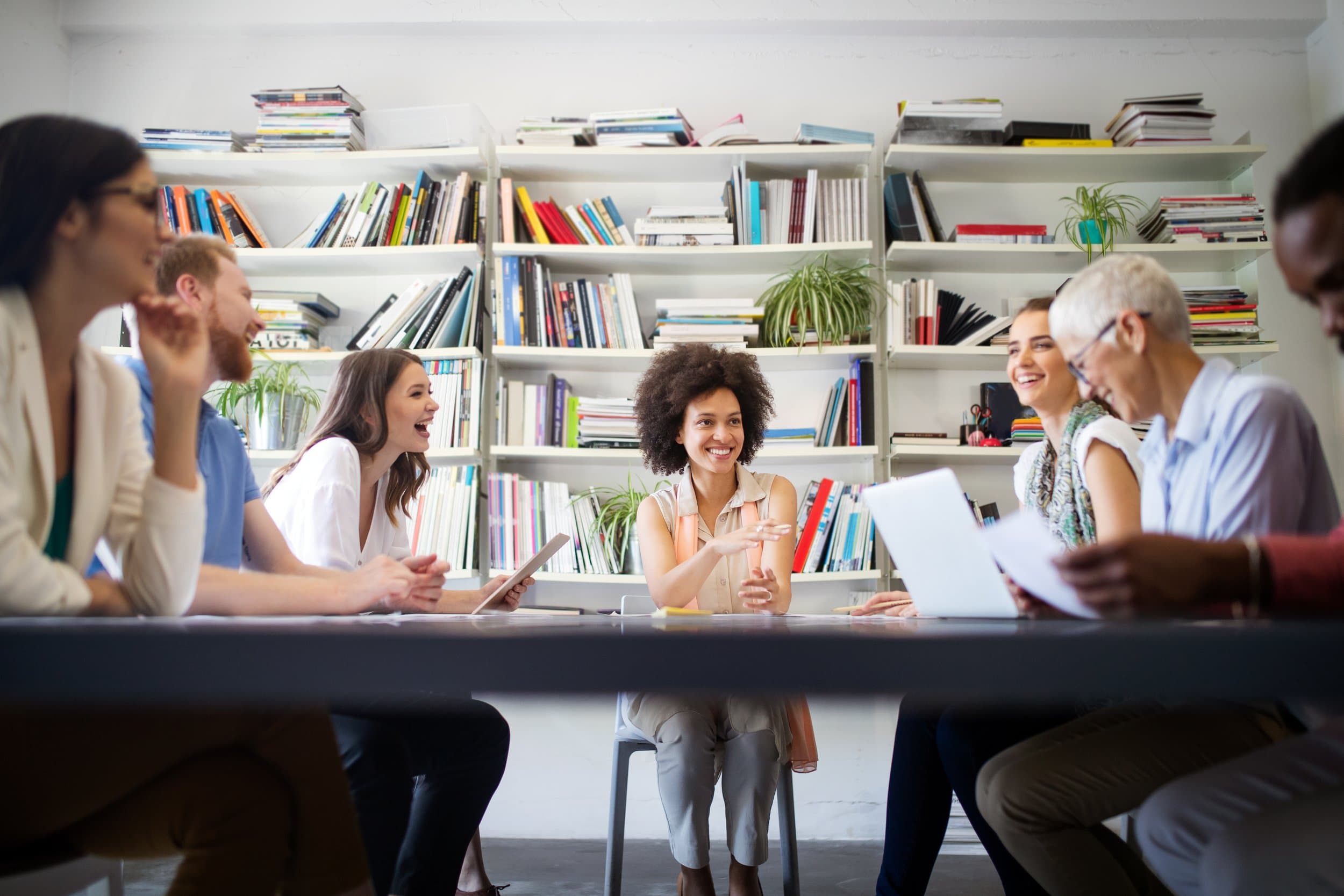 A group of adults laugh and talk around a long table with a tablet and laptop, in front of tall shelves of books.