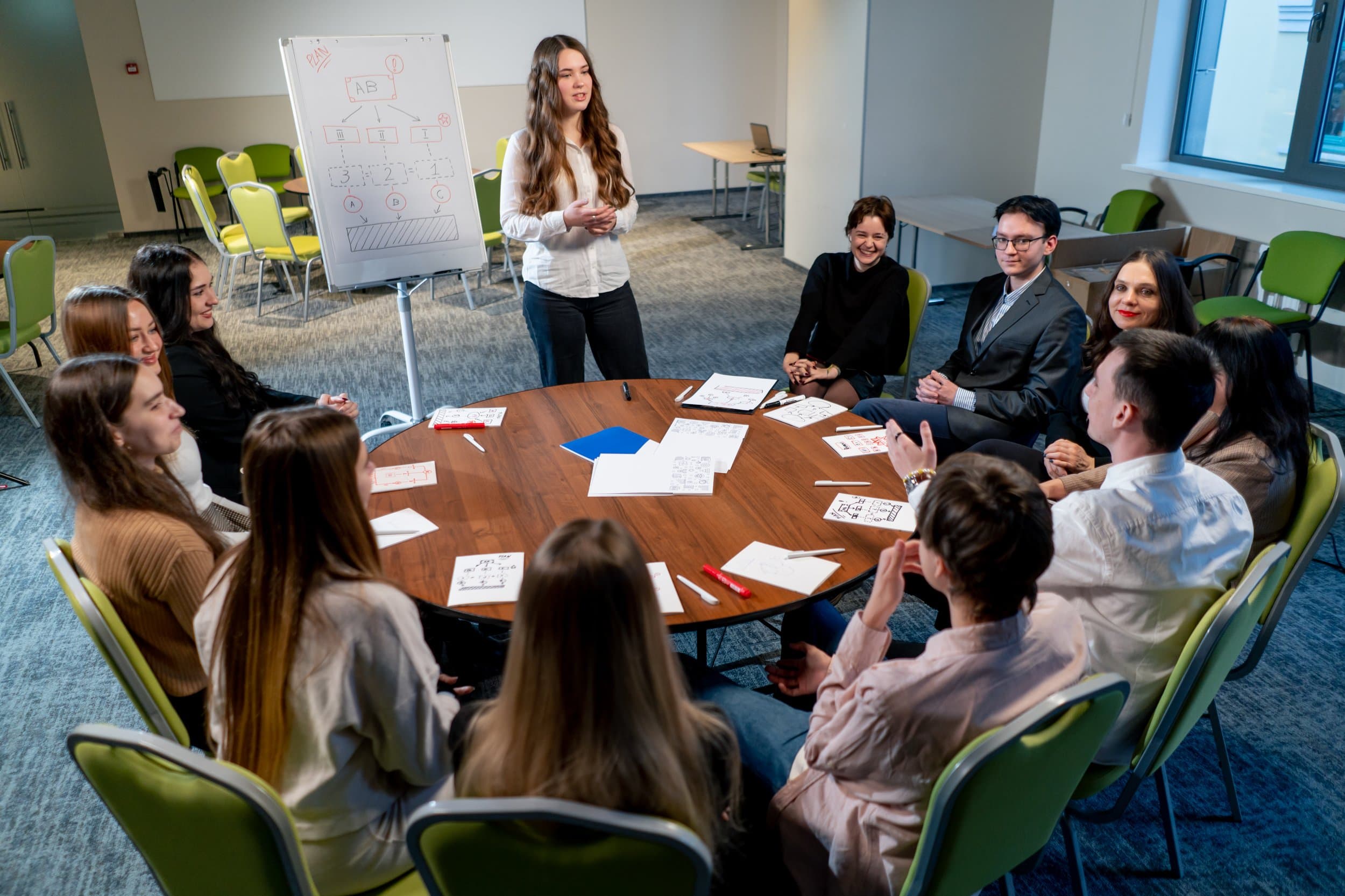 A person stands beside a flip chart diagram and speaks to a group of adults seated around a round table.