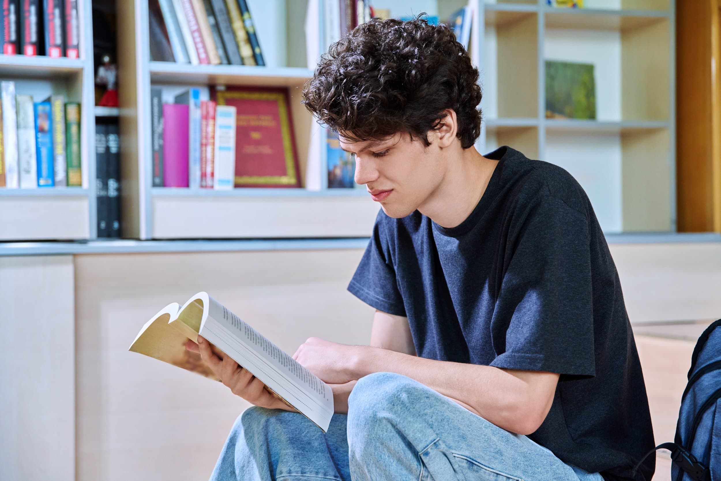 A student sits on a library floor reading a thick paperback book in front of shelves of books.