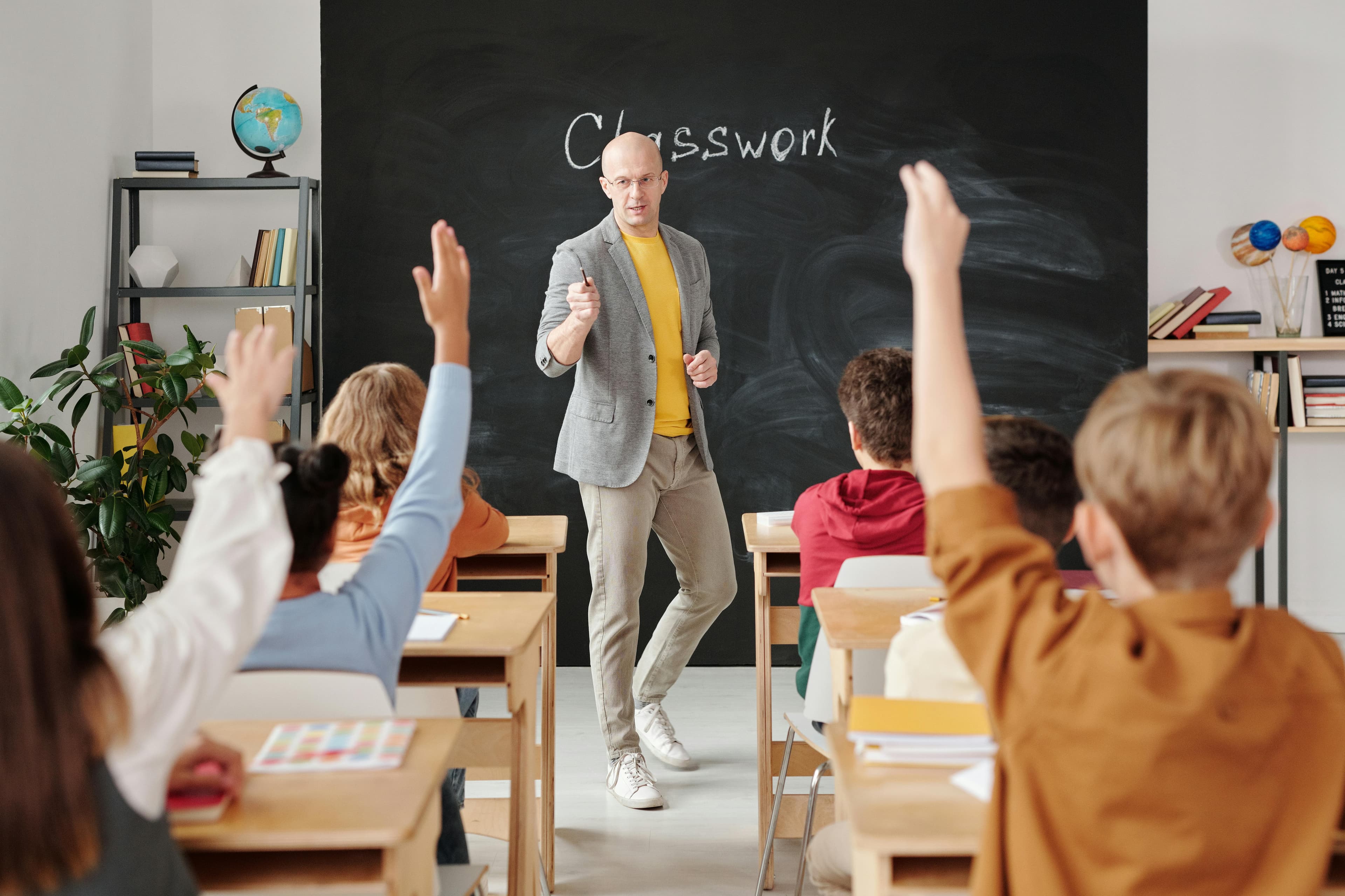 A teacher at a chalkboard with students engaged and raising their hands.